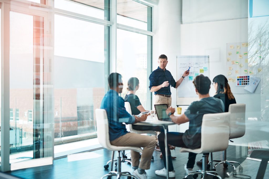 A man presenting a business strategy to a diverse group of colleagues in a modern, brightly lit office meeting room.