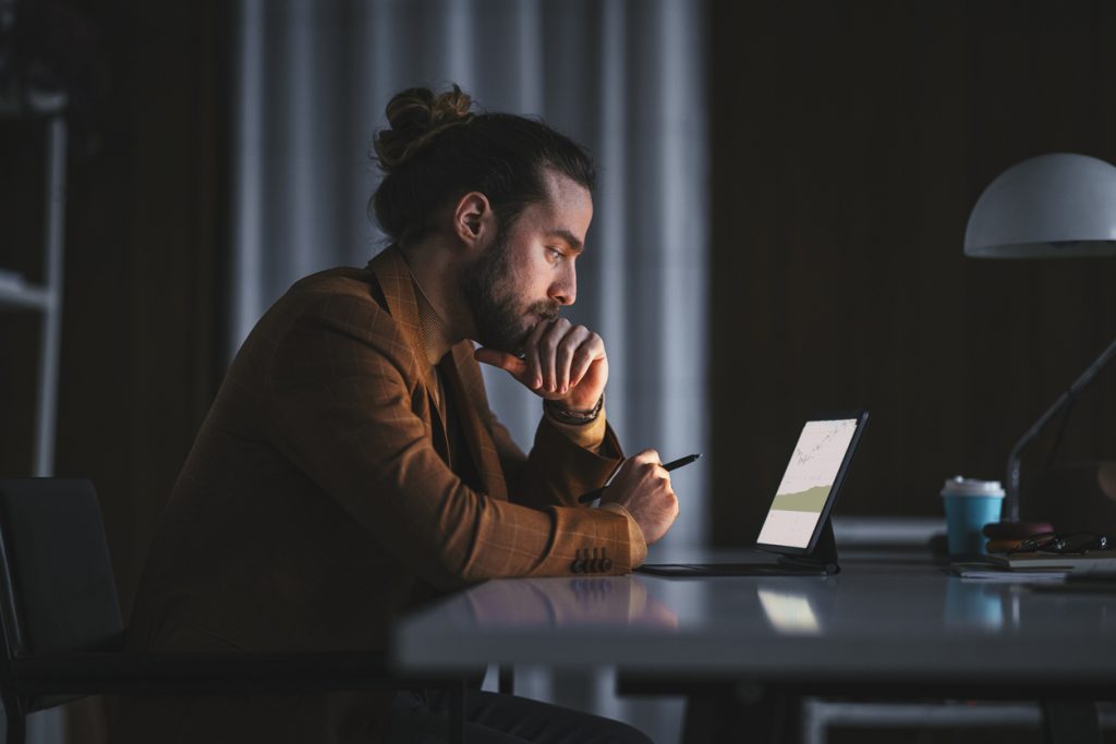 Man with a bun working intently at his desk in a dimly lit room, using a tablet and stylus with a coffee cup nearby.