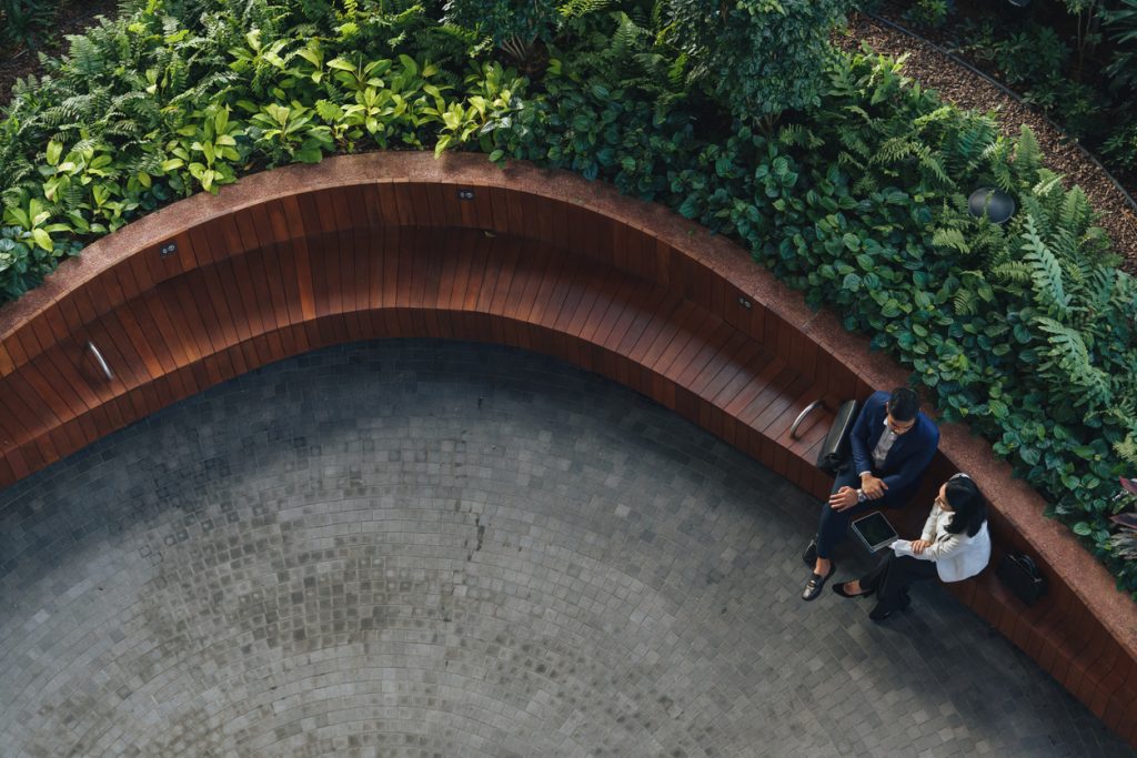 Two people sitting and talking in a curved wooden seating area surrounded by lush green plants, viewed from above.