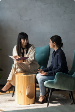 Two women engaged in a conversation, one holding a document, sitting in a modern office with stylish chairs and a wooden table.