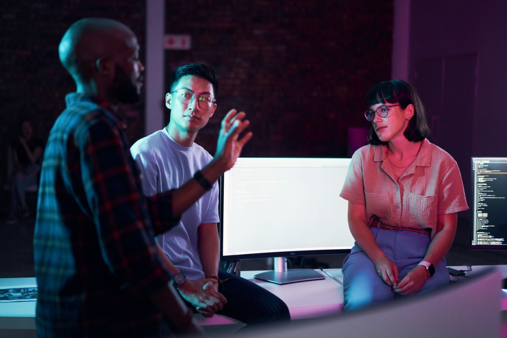 Three colleagues discuss a project in a dimly lit office, with one explaining a point as they stand beside computer monitors displaying code.