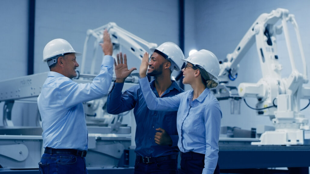 Three industrial engineers in hard hats discussing operations in a factory with robotic arms in the background.