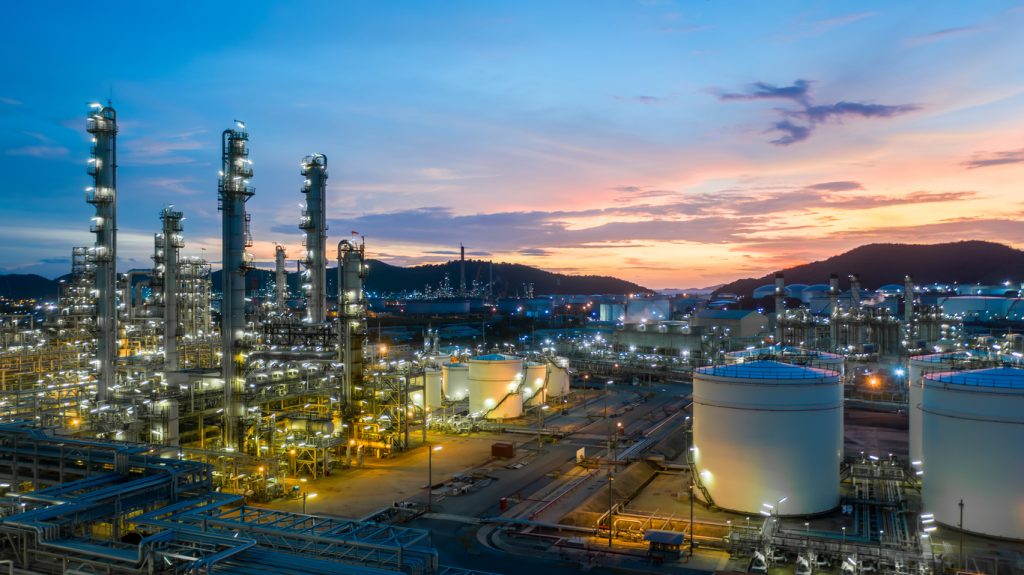 An industrial refinery complex with large storage tanks and tall structures is illuminated at dusk, with mountains and a colorful sky in the background.